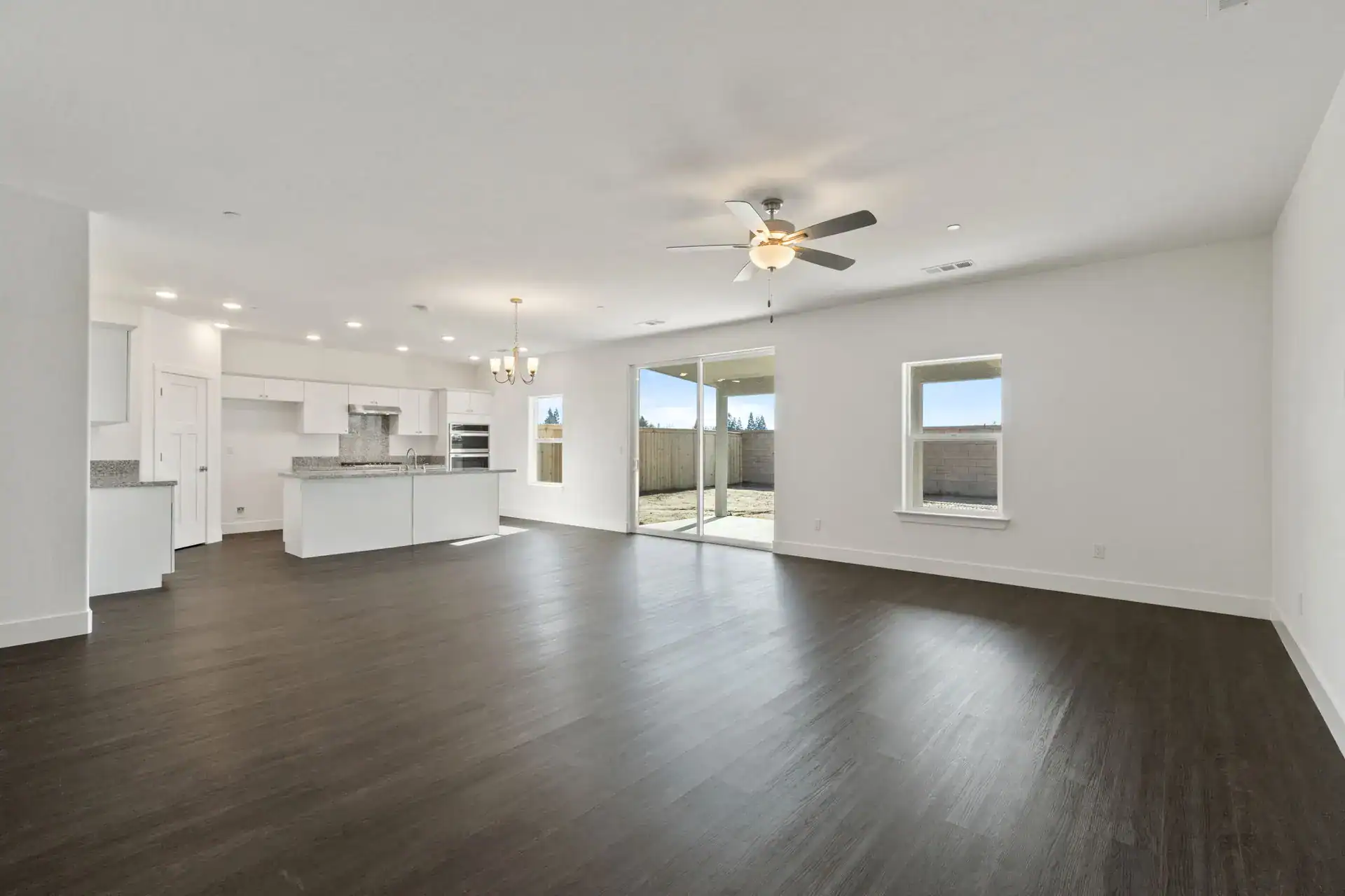 Spacious living area with white walls, dark flooring, ceiling fan, and ample natural light from larg.